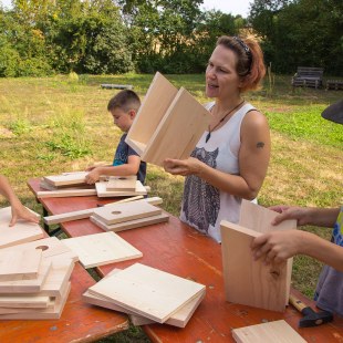 Janina Kempf steht an einem Tisch, auf dem Bretter für Nistkästen liegen. Sie hält einen halbfertigen Kasten in der Hand und redet, auf dem Bild sind noch drei Kinder zu sehen, die ebenfalls bauen | © memo AG