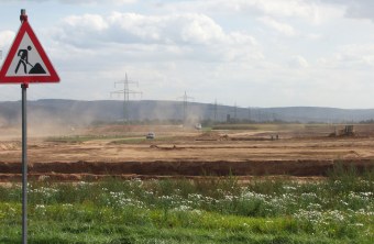 Grüne Wiese im Vordergrund, dahinter eine planierte Fläche mit Autos und Traktoren. Links steht ein Baustellenschild | © Thomas Staab