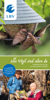 Eine Gruppe von Vögeln versammelt sich an einem Futterhaus, während zwei Personen im Freien den LBV-Flyer betrachten.