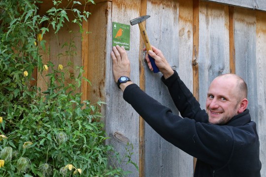 Ein Mann lacht in die Kamera und hält in der rechten Hand einen Hammer und in der linken die Plakette "Vogelfreundlicher Garten" | © Carola Bria