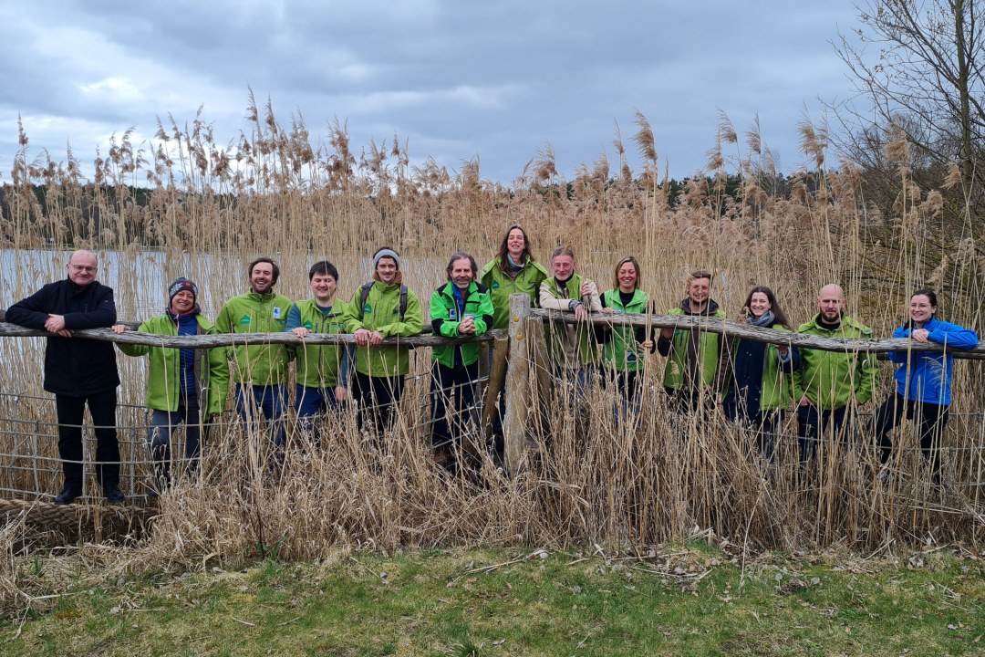 Ganz links steht LBV-Vorsitzender Dr. Norbert Schäffer, daneben 13 Gebietsbetreuer und -betreuerinnen in grünen Jacken, sie stehen an einem See hinter einem Holzzaun. Dahinter ist Schilf. | © LBV