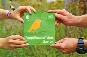Zwei Hände halten ein grünes LBV-Schild mit der Aufschrift "Vogelfreundlicher Garten" in einem blühenden Garten.