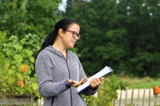 Eine Frau mit Brille steht seitlich, blickt nach rechts und spricht mit Klemmbrett in der einen Hand und Stift in der anderen | © Carola Bria