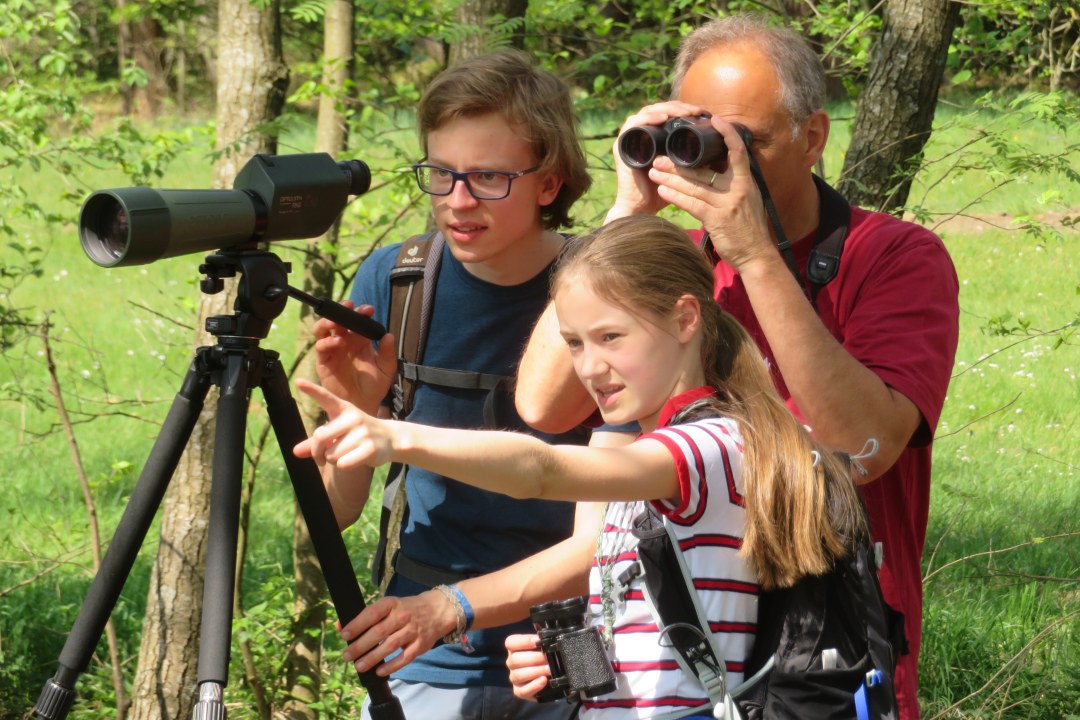 Zwei Kinder deuten in eine Richtung, vor Ihnen ein Spektiv auf einem Stativ, im Hintergrund ein Erwachsener, der durch ein Fernglas sieht | © Jakob Braun