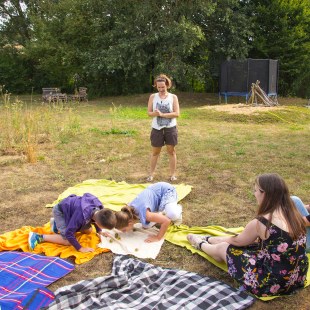 Janina Kempf und vier Kinder sitzen auf einer Wiese, die Kinder auf Decken. Janina steht und erklärt etwas, zwei Kinder spielen ein Spiel | © memo AG