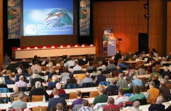Großer Saal mit Leinwandprojektion und rechts einem Pult, wo Ulrike Scharf steht. Auf Bänken sitzen viele Menschen und schauen nach vorn | © Claudia Becher