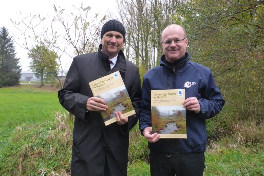 Dr. Uwe Brandl (Bayerischer Gemeindetag) und Dr. Norbert Schäffer (LBV) stehen im Grünen und zeigen die Praxisbroschüre "Lebendige Bäche in Bayern" | © Ralf Hotzy