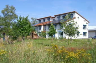 Weißes Gebäude mit blauen Fenstern der Landesgeschäftsstelle in Hilpoltstein. Dafür ist ein grüner Garten zu sehen mit Bäumen, einem Geräteschuppen und einigen Sträuchern | © R. Brode