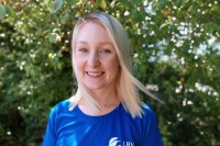 Eine lächelnde Frau mit blonden Haaren trägt ein LBV-T-Shirt im Freien, umgeben von grünen Blättern.