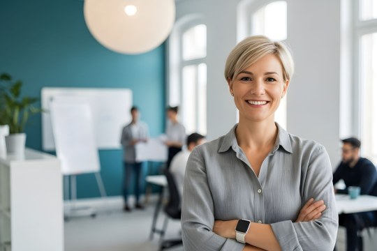 Eine lächelnde Frau steht mit verschränkten Armen in einem modernen Büro, während im Hintergrund ein Meeting stattfindet. | © stock.adobe.com