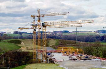 Kräne stehen auf einer Baustelle im Grünen, wo eine Brücke gebaut wird. | © Andreas Hartl