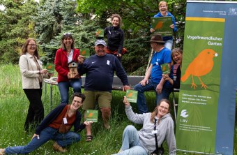 Gruppe von Menschen hält LBV-Schilder in einem grünen Garten, umgeben von Bäumen, lachend und posierend.