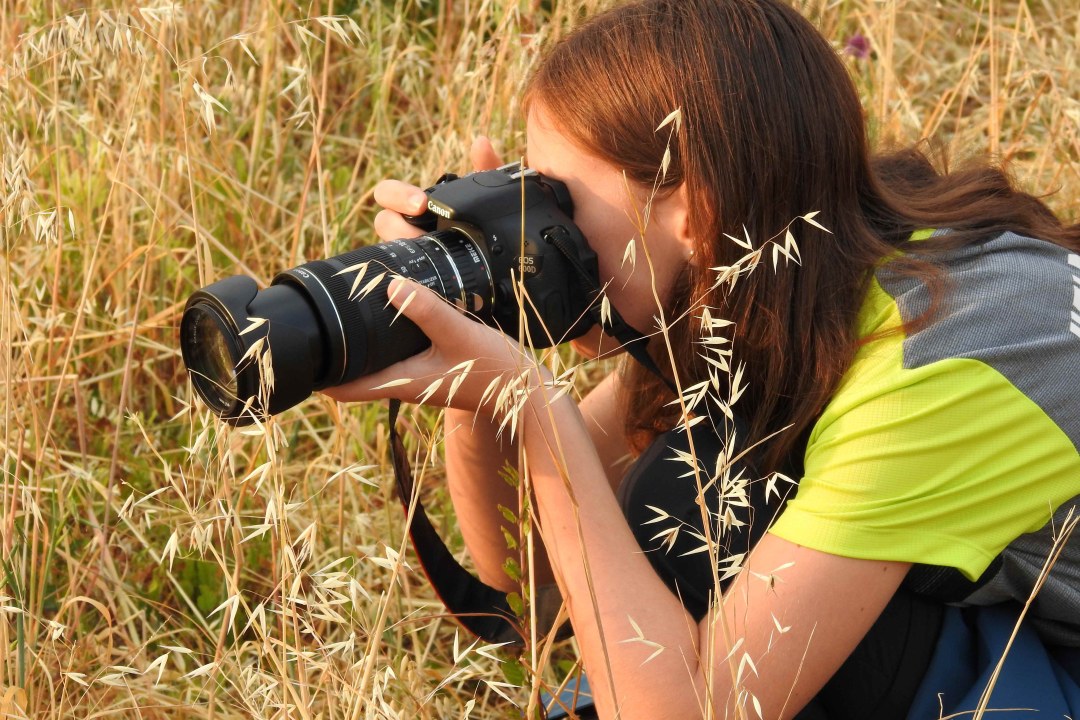 Kind sitzt in einem Feld und fotografiert mit einer Kamera. | © Andrea Koch