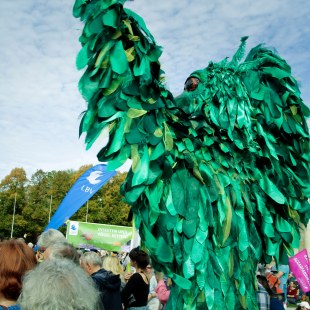 Stelzentänzer als grüner Vogel verkleidet auf der Demo Mia ham's satt aus einer leichten Froschperspektive von schräg hinten fotografiert | © LBV