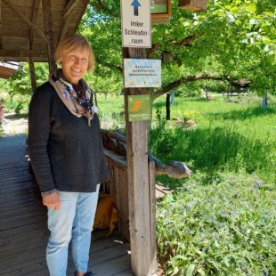 Eine Frau steht lächelnd neben einem Schild zu einem Imker-Schleuderraum im LBV-Naturschutzgarten.