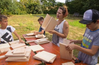 Janina Kempf steht an einem Tisch, auf dem Bretter für Nistkästen liegen. Sie hält einen halbfertigen Kasten in der Hand und redet, auf dem Bild sind noch drei Kinder zu sehen, die ebenfalls bauen | © memo AG